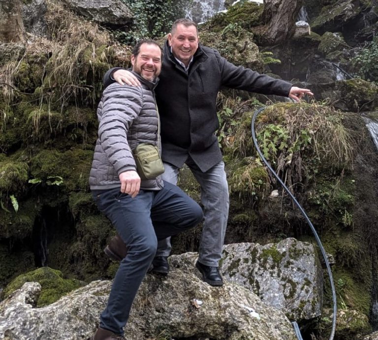 Zwei Personen posieren lächelnd vor einem Wasserfall in einer natürlichen Umgebung.