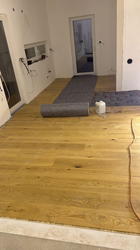 Holzboden mit unvollständiger Verlegung, graue Teppichrolle im Raum.