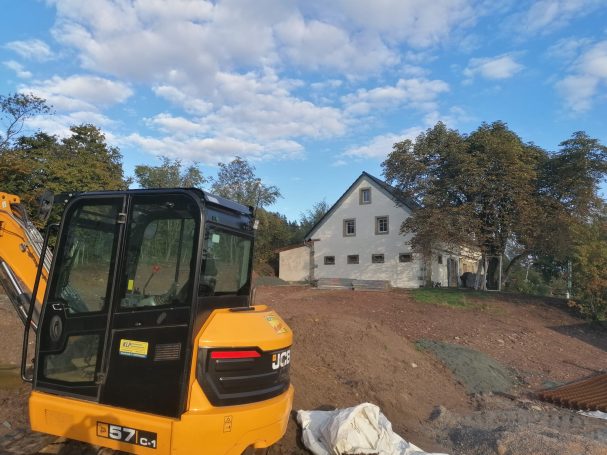 Baustelle mit Gelbbagger und einem unvollendeten weißen Haus im Hintergrund.