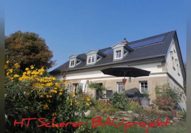 Zweifamilienhaus mit Dachgauben und Solarzellen, umgeben von Blumen und Bäumen.