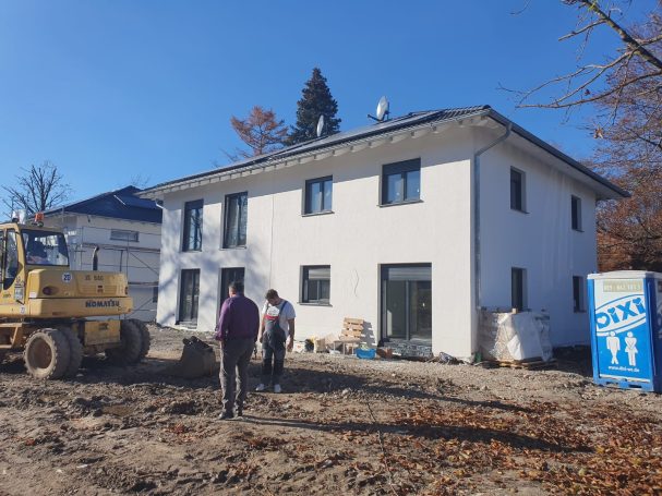 Bauarbeiter stehen vor einem unvollendeten, weißen Wohnhaus in einer Holzbaulandschaft.