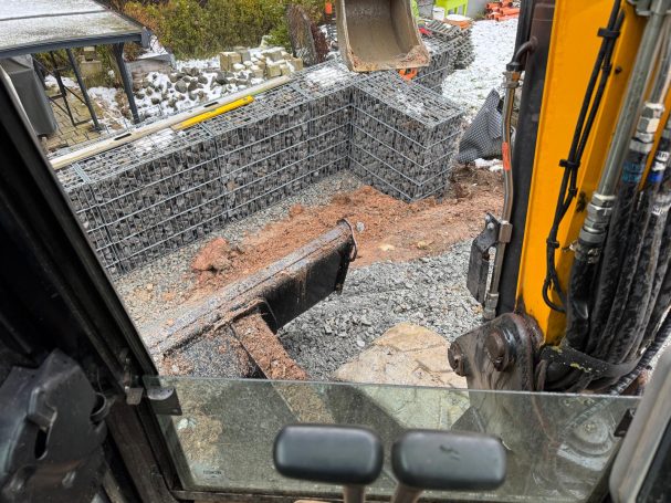 Ralf Scherer sitzt im Bagger und setzt mit Mitarbeiter Gabionen