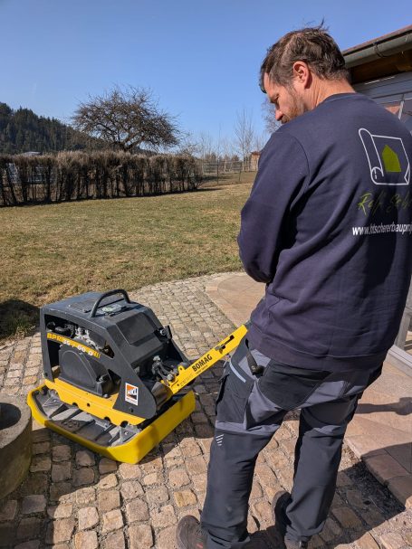 Ein Mann bedient eine Rüttelplatte auf einem gepflasterten Außenbereich.