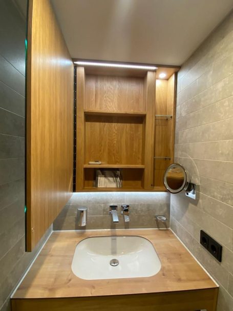 Modern Badezimmer mit Holzregal, Waschbecken und Spiegelbeleuchtung.